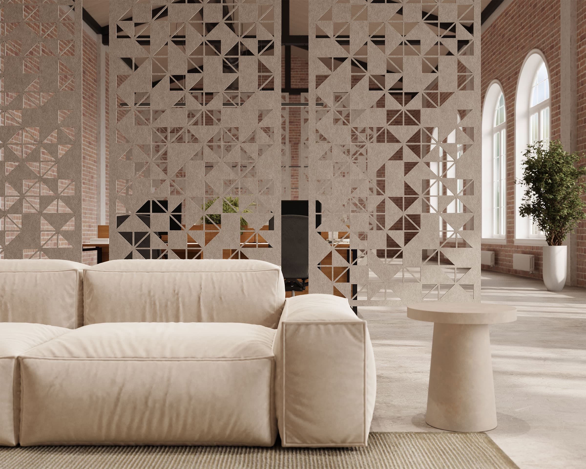 A modern, minimalist living room featuring a beige sofa and a small matching side table. Behind the sofa, decorative geometric partitions and acoustic felt screens partially separate the space. The room is brightened by arched windows and includes a potted plant.