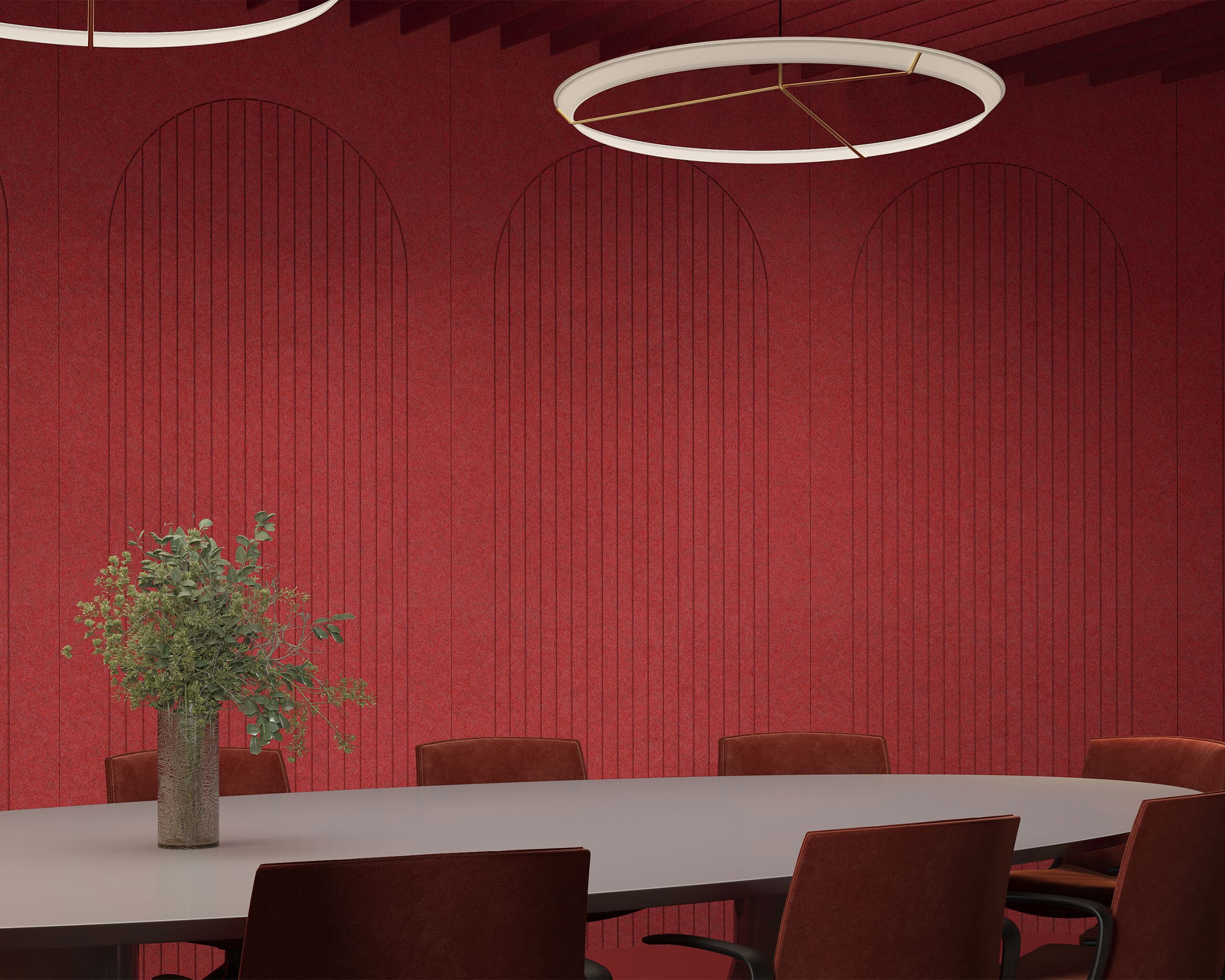 A modern conference room features a large circular table surrounded by brown chairs. A vase with green foliage sits at the center of the table. The red walls, adorned with vertical line patterns and arched detailing, are complemented by arch acoustic felt wall panels. Two circular light fixtures hang from the ceiling.