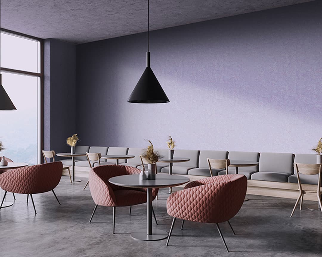 A modern café interior with lavender acoustic felt wall panels and dark gray flooring. The space features round tables with pink, quilted chairs and light wood chairs with gray cushions against the wall. Acoustic felt wall panels add a touch of sophistication, while minimalist black pendant lights hang from the ceiling and dried flowers adorn the tables.