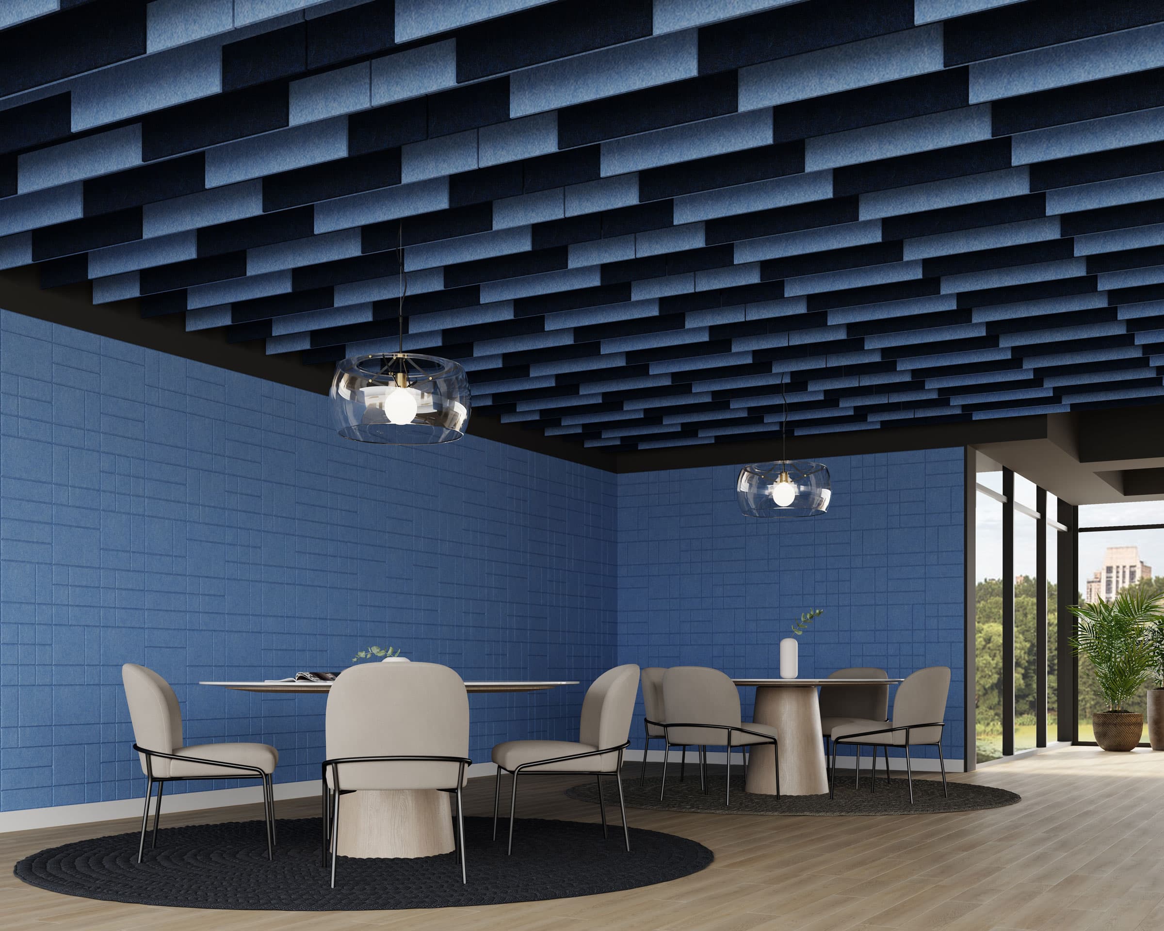 A modern interior features a meeting area with beige chairs around circular tables on black rugs. The room has blue walls and a ceiling with geometric blue acoustic felt ceiling baffles. Two pendant lights with transparent shades hang above the tables, and large windows offer a view of greenery outside.