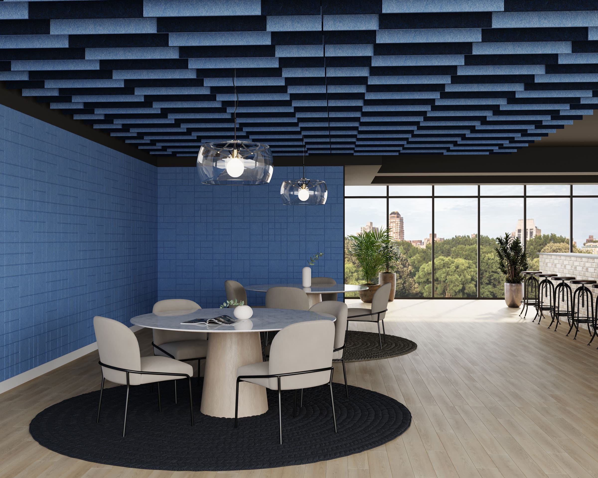 A modern office space features a round marble table with cream-colored chairs on a black circular rug. Above, there are artistic blue acoustic felt ceiling baffles and pendant lights. Floor-to-ceiling windows offer a view of a city skyline and greenery.