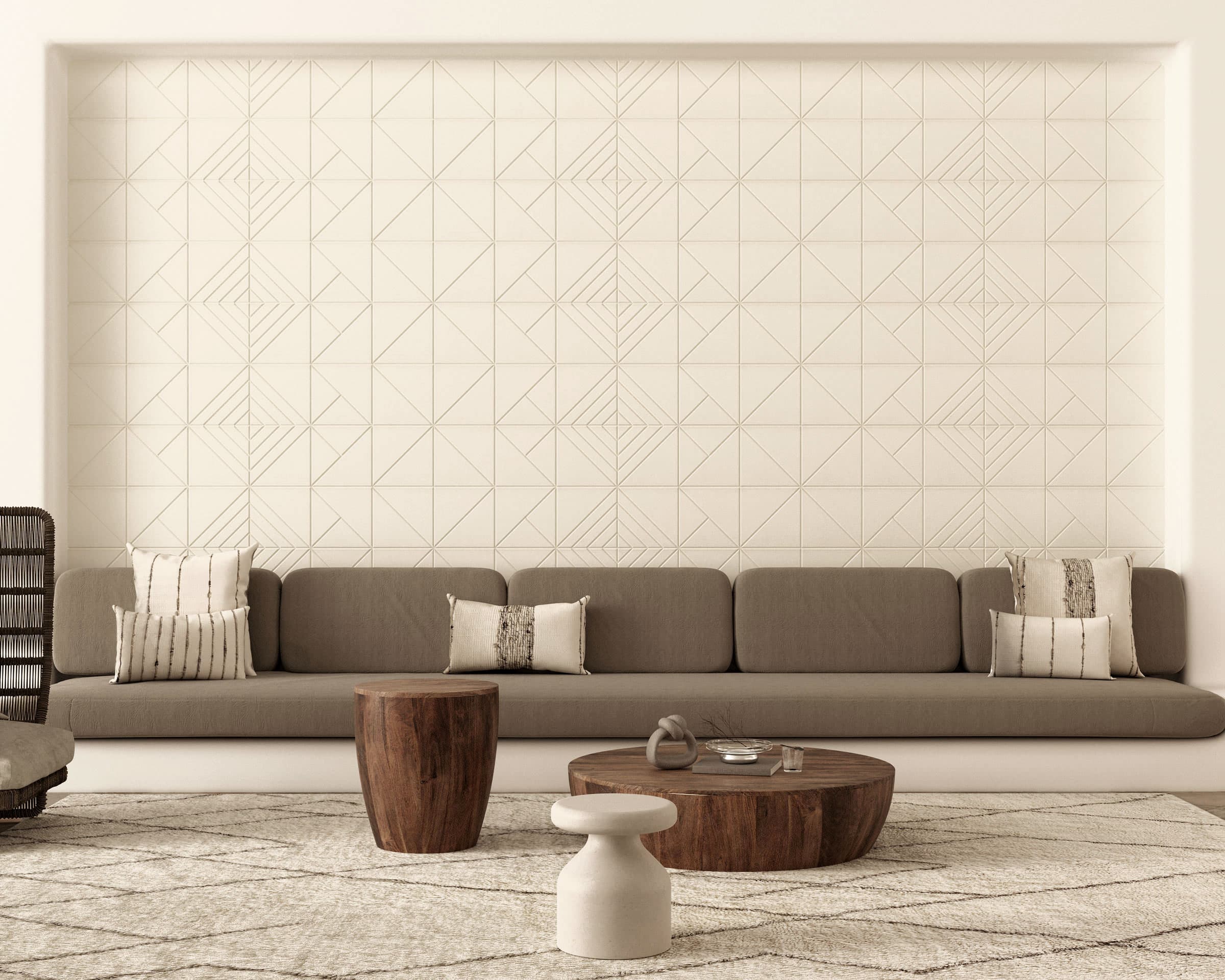 A modern living room with a long gray sofa adorned with patterned cushions against a geometric-patterned white wall featuring acoustic felt wall tiles. In front of the sofa are three wooden and stone coffee tables of varying heights on a textured beige rug.