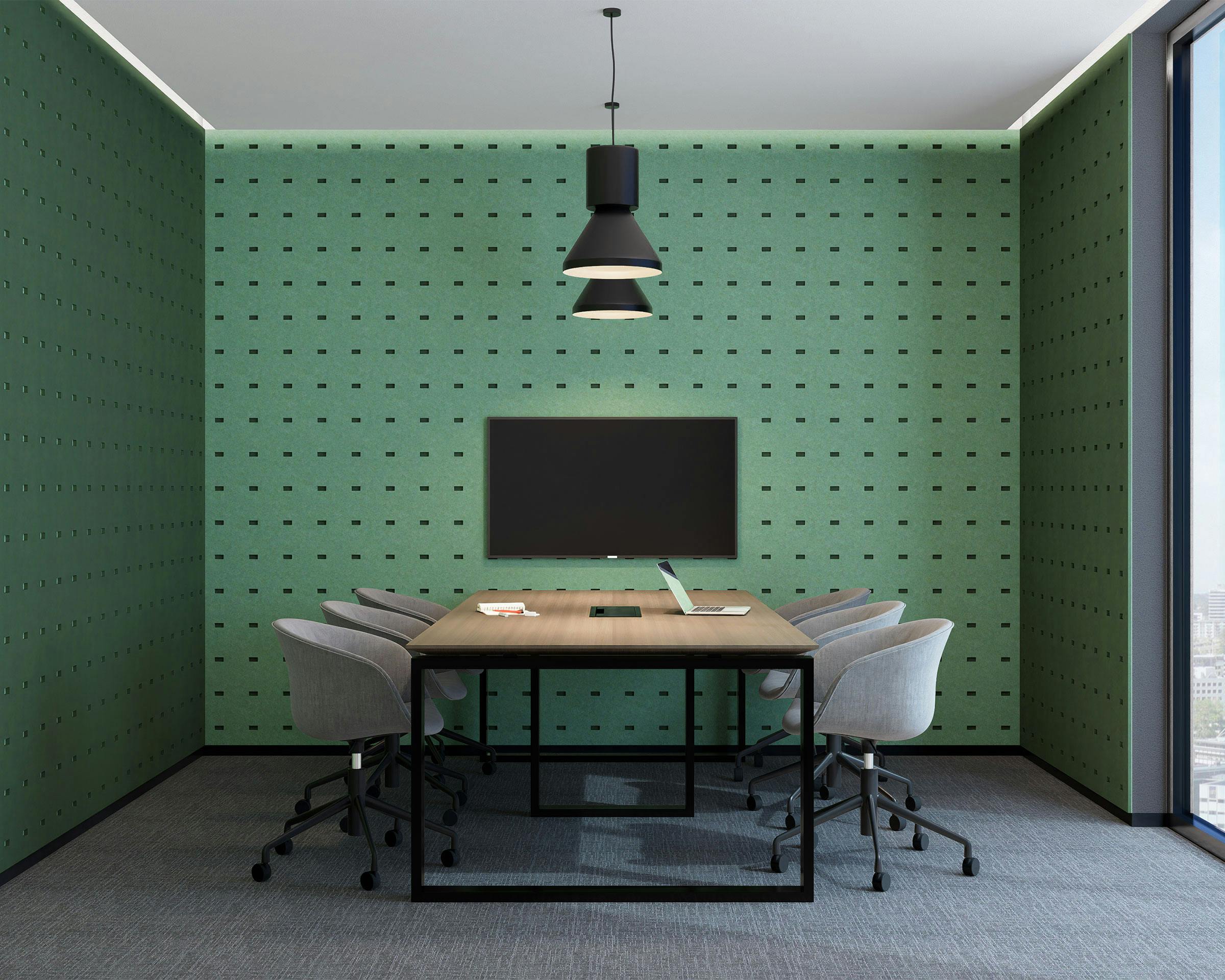 A modern conference room with a long wooden table and six gray chairs on casters. The room features a green acoustic felt wall panels with a pattern of small rectangles for sound absorption, a ceiling light fixture, a large flat-screen TV or monitor on the wall, and a large window with a city view.