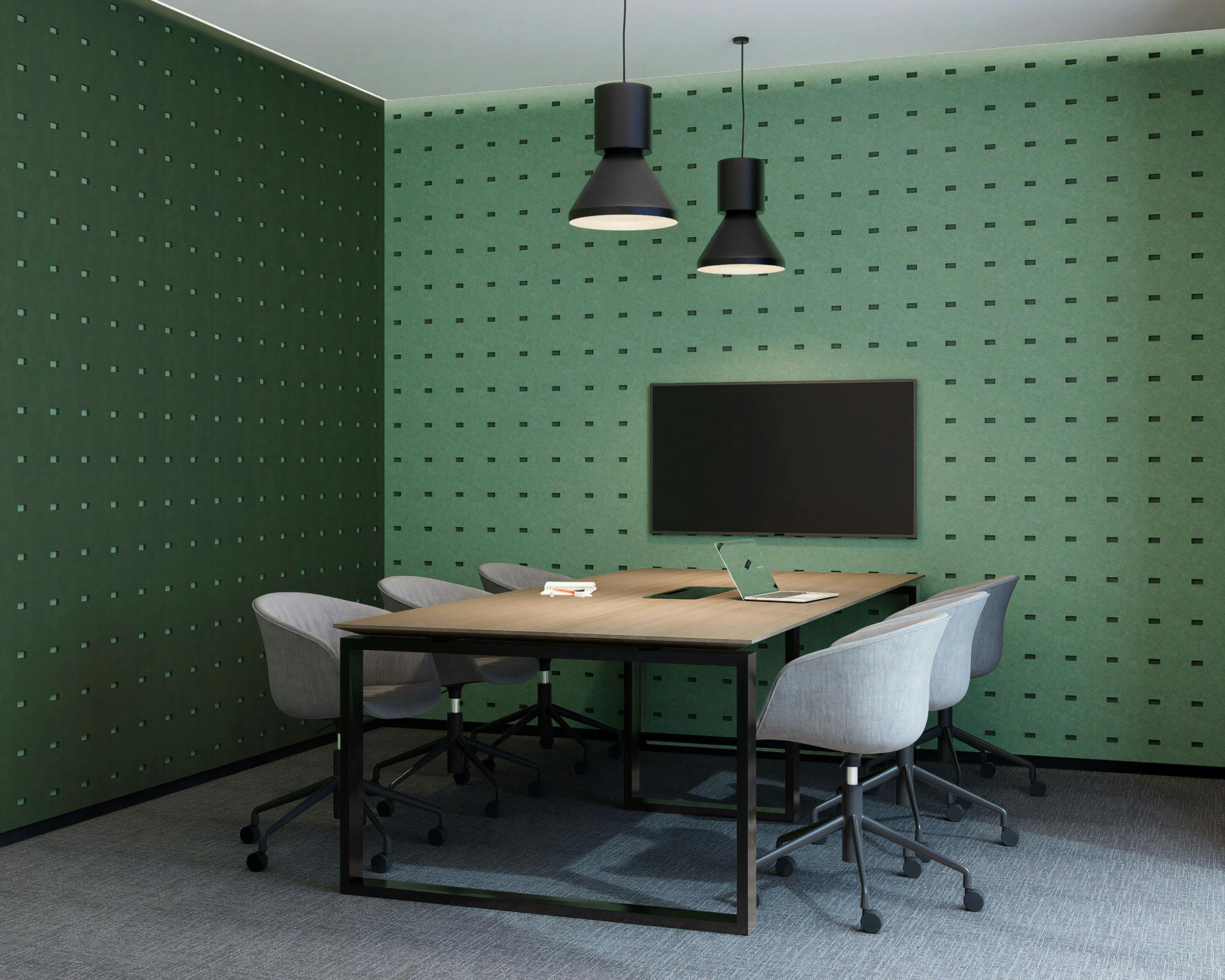 A modern conference room features a wooden table surrounded by six gray office chairs on wheels. A flat-screen TV is mounted on a green wall lined with rectangle acoustic felt wall panels. Two black pendant lights hang over the table, and a green laptop is placed on the table.