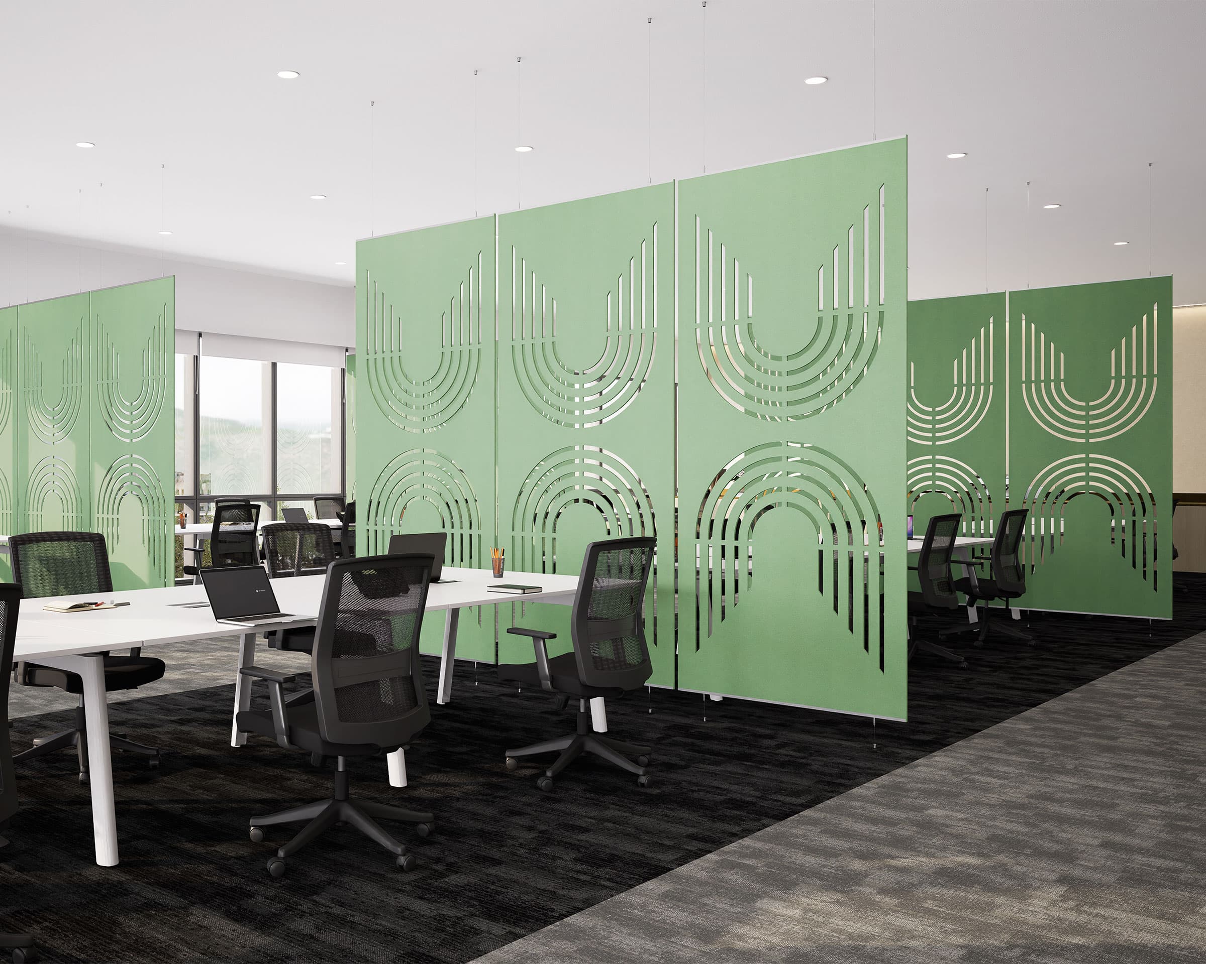 An open-plan office space with modern furniture, featuring several workstations equipped with chairs and laptops. Green acoustic felt panels with geometric cut-out designs separate the desks, while large windows allow natural light to fill the room.