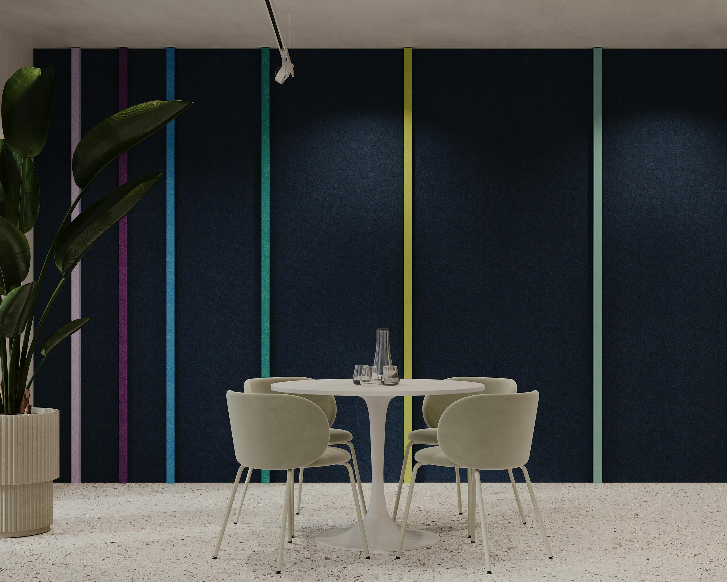 A modern, minimalist interior features a small white round table with four light green chairs. The backdrop is a dark blue acoustic felt wall accented with vertical, colorful stripes that resemble finely stitched seams. A potted plant with large green leaves stands on the left, adding a touch of nature.