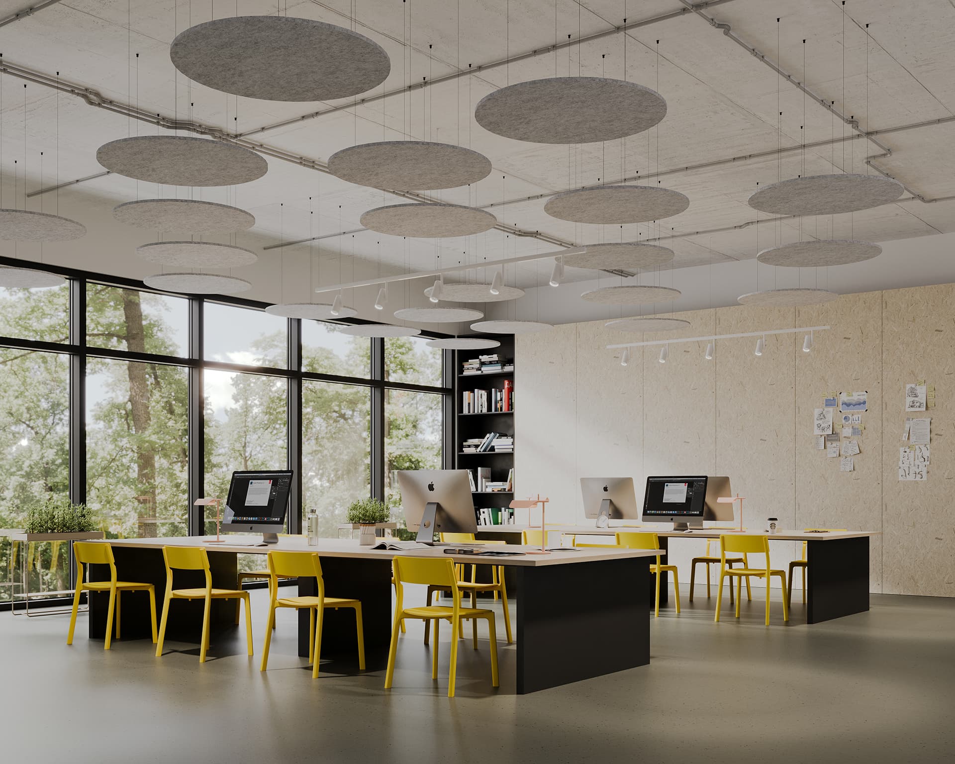 A modern, open-concept office space featuring long shared desks with desktop computers and yellow chairs. Large windows line the wall, offering a view of trees outside. Circle acoustic felt ceiling clouds are suspended from the ceiling, and bookshelves line the back.