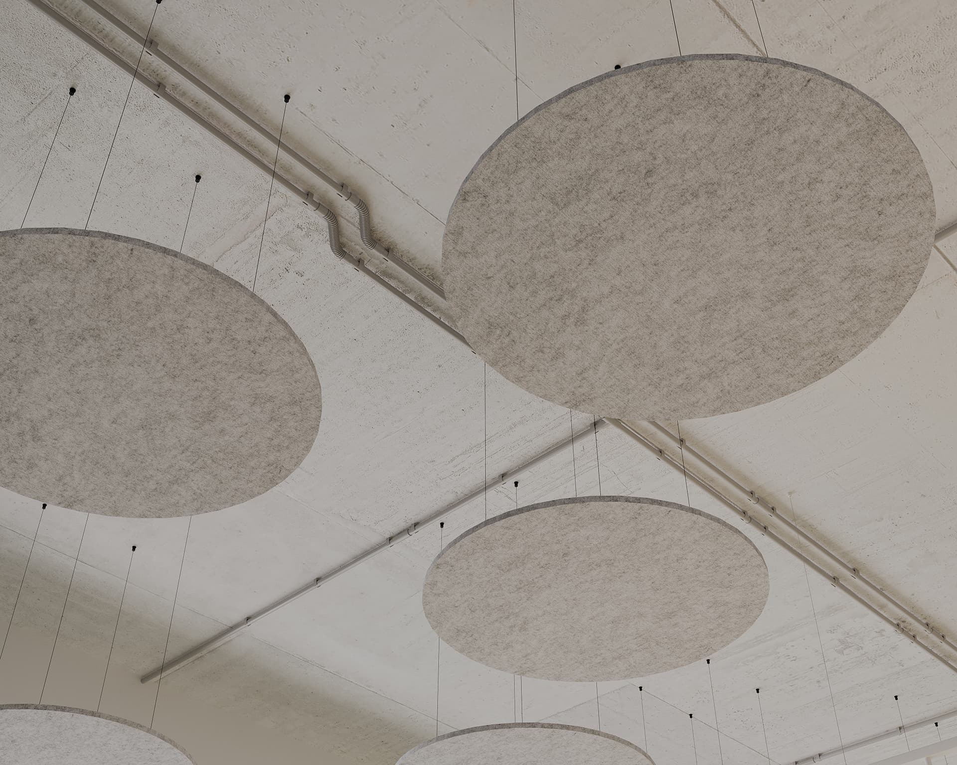 Circular gray acoustic felt clouds are suspended from the ceiling by thin wires in a minimalist, industrial-style room. The ceiling has exposed piping, enhancing the modern aesthetic and functionality.