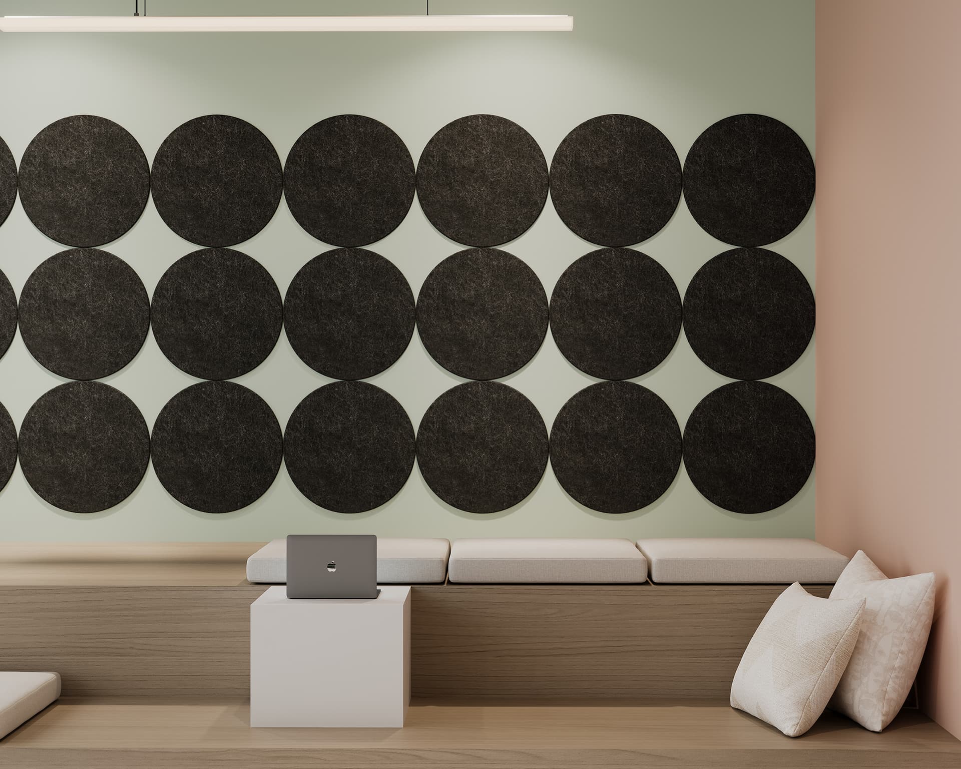 A modern interior features a light green wall with a grid of 15 large black circular acoustic tiles. Below, there are beige cushioned benches with a white pillow and a laptop placed on a small white table. The room is softly lit with a minimalistic light fixture.