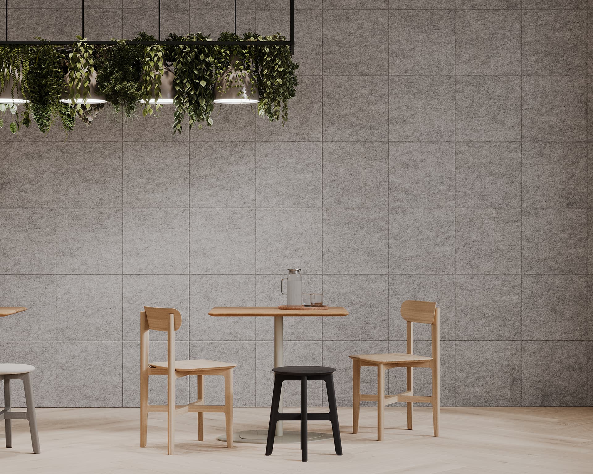 Minimalist cafe interior featuring a small, round wooden table with three mismatched chairs and a black stool. A tall glass bottle with a wooden stopper is on the table. The gray acoustic tiled wall in the background has a decorative hanging plant display above.