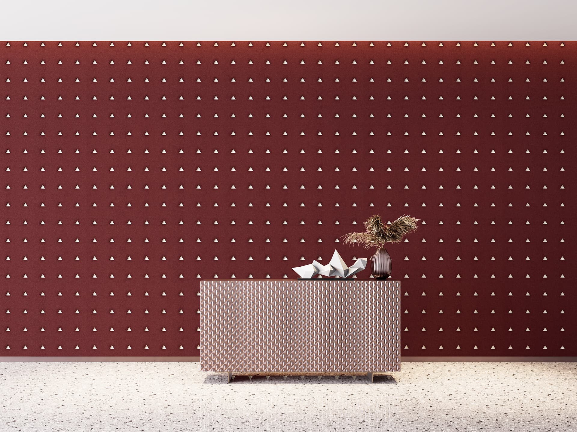 A modern interior with a maroon acoustic felt wall featuring a repeating triangular pattern. In front, a textured beige table holds a minimalist origami sculpture and a clear vase with dried flowers. The floor is a light speckled terrazzo.