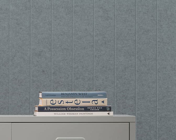 A minimalist scene with four essential books stacked on a gray cabinet against a gray textured acoustic felt wall. The books are titled "Benjamin West," "A Possession Obsession," and "William Wegman Paintings," along with one book whose title is partially obscured.