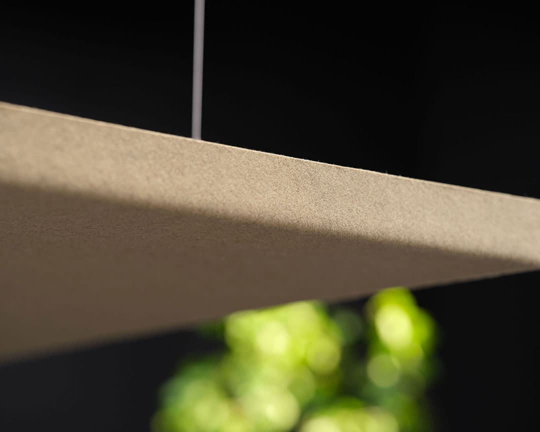 A close-up shot of a beige, rectangular suspended acoustic felt ceiling panel against a dark background. The panel has a smooth texture and is hanging via thin, almost invisible wires. Out of focus, vibrant green foliage can be seen in the background, adding a touch of naturals to the scene.