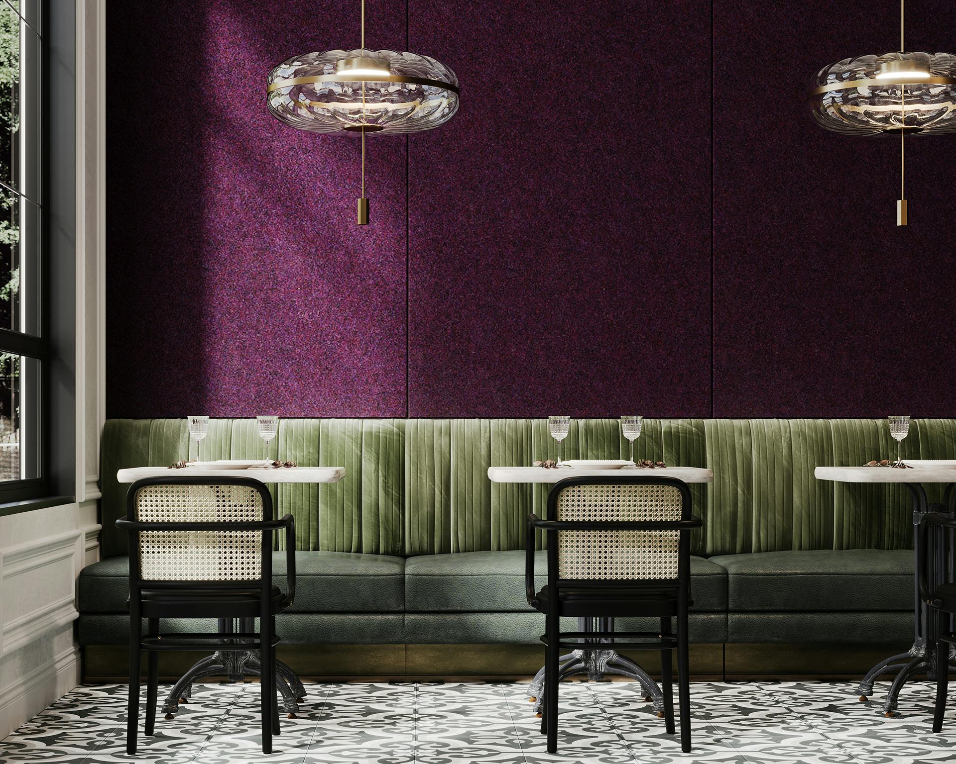 A chic restaurant interior featuring green velvet banquette seating against a dark purple textured acoustic felt paneled wall. Black and white patterned tile floor complements the space. Three modern pendant lights hang above marble-topped tables with black chairs.