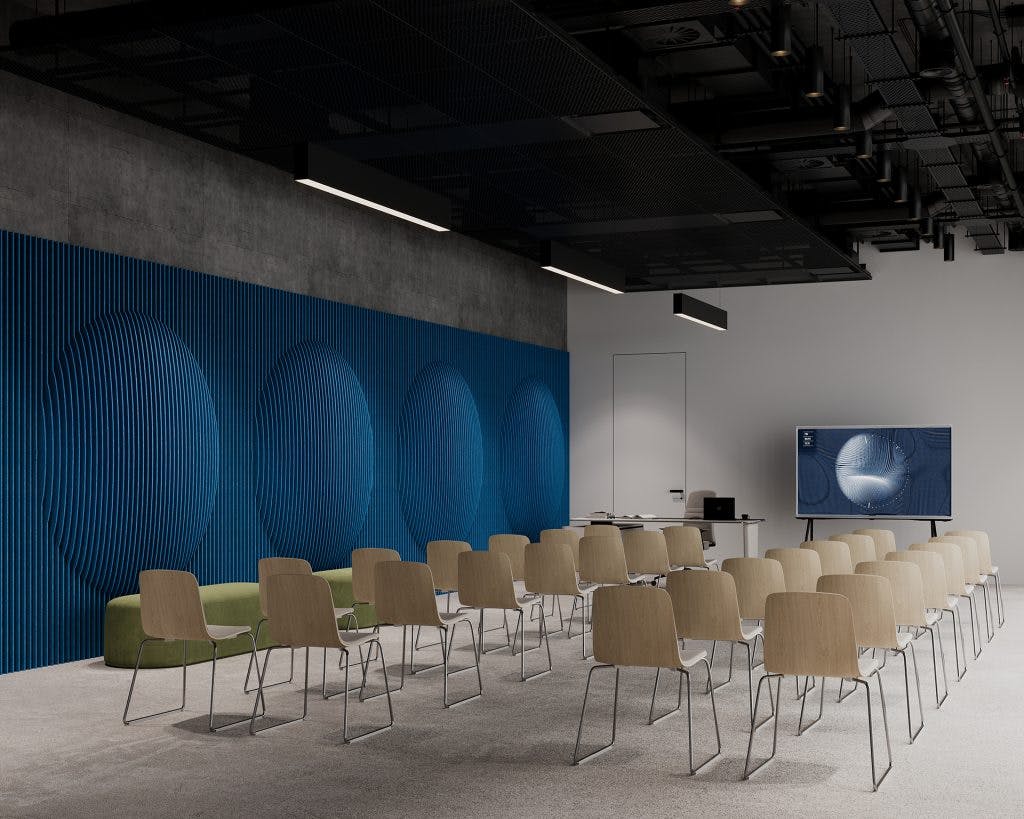A modern conference room with rows of beige chairs facing a large screen on the right. The room features a blue textured wall with circular patterns, a green bench, and a ceiling with exposed pipes and suspended lights. A door is visible at the back.