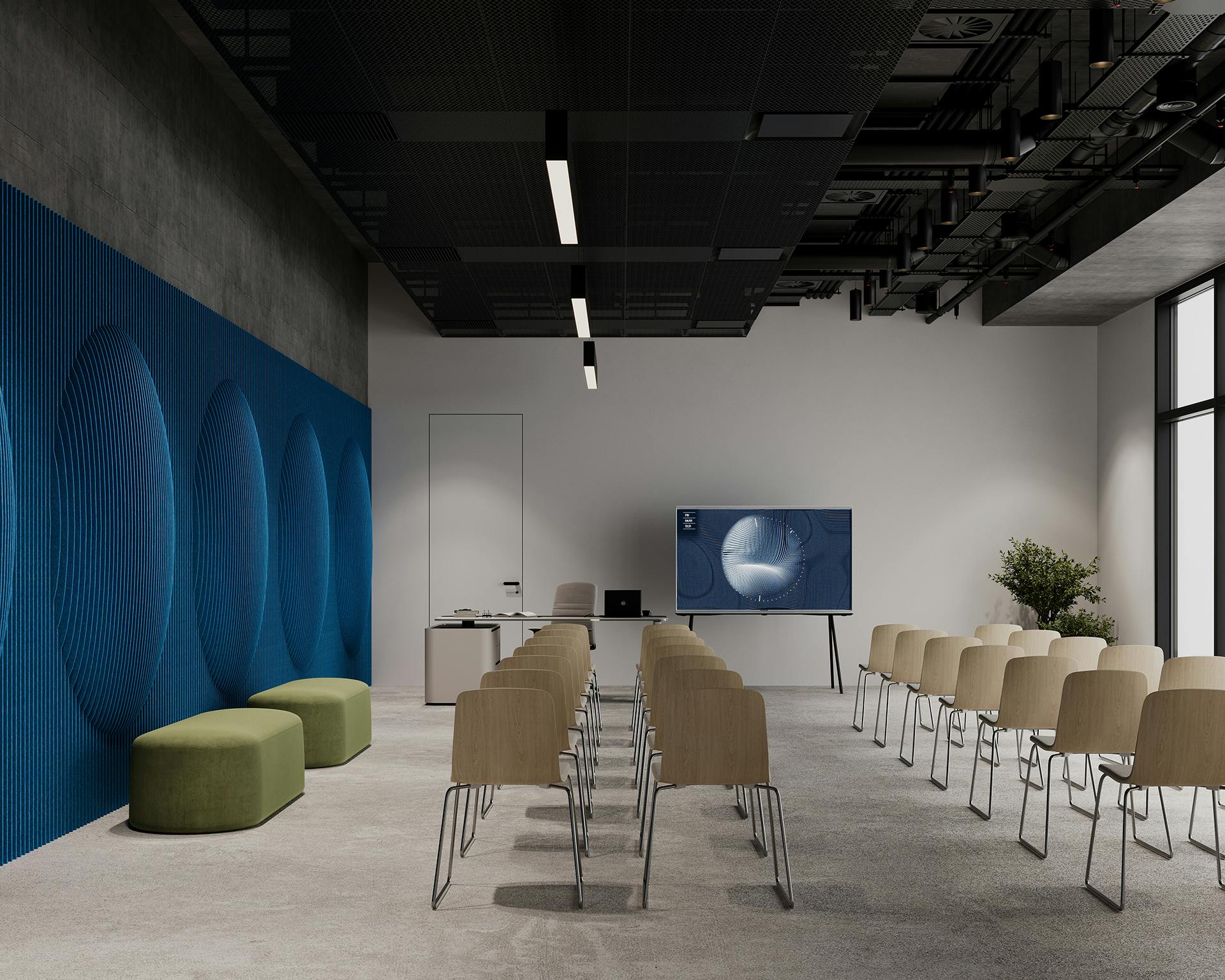 A modern conference room features rows of wooden chairs facing a screen displaying a presentation. The room has a minimalist design with a blue textured wall, green ottomans, and a desk at the front. Large windows allow natural light to fill the space.