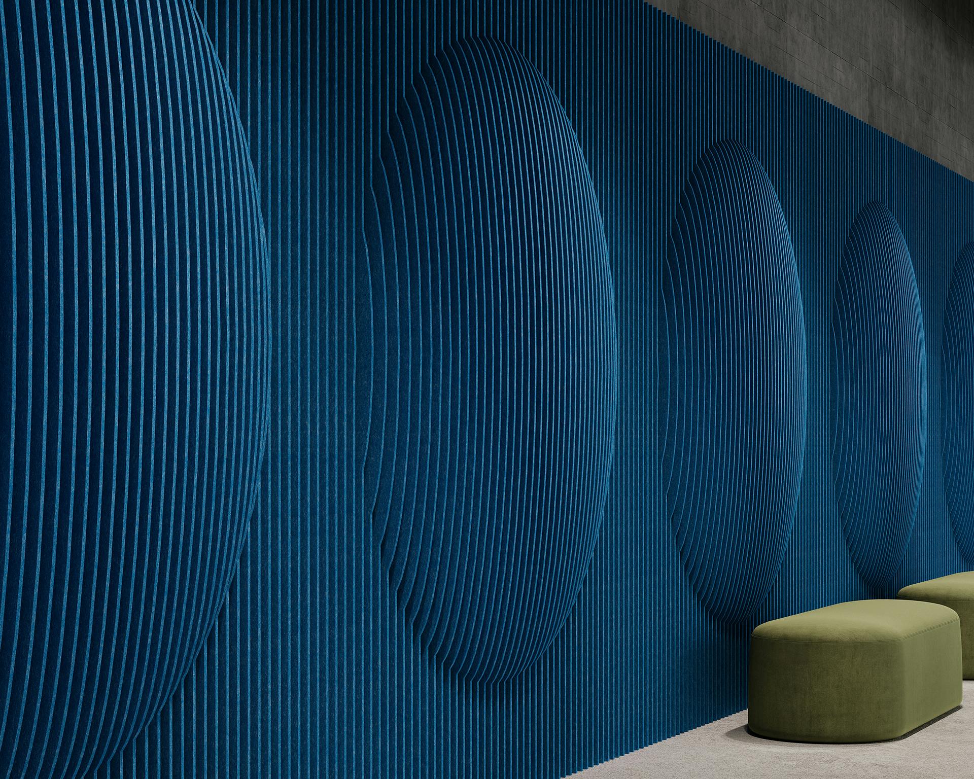 A modern wall art installation featuring a series of large, indented oval shapes on a textured, ribbed blue surface. A single olive-green cushioned bench is placed against the wall on a light gray floor. The overall scene is minimalist and contemporary.