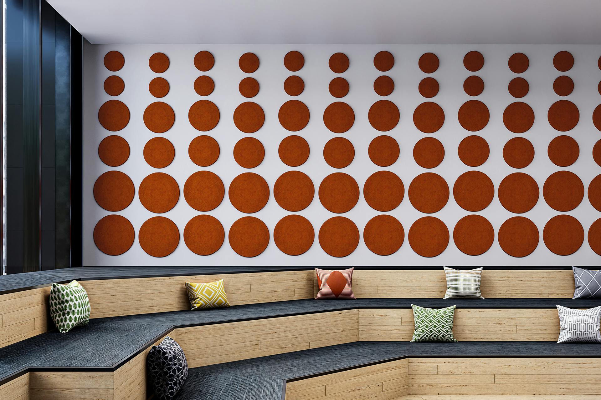 A modern seating area with multi-tiered wooden platforms and scattered decorative pillows in various patterns. The white wall in the background features an artistic arrangement of orange circular acoustic felt panels in a grid pattern. Floor-to-ceiling windows are to the left.