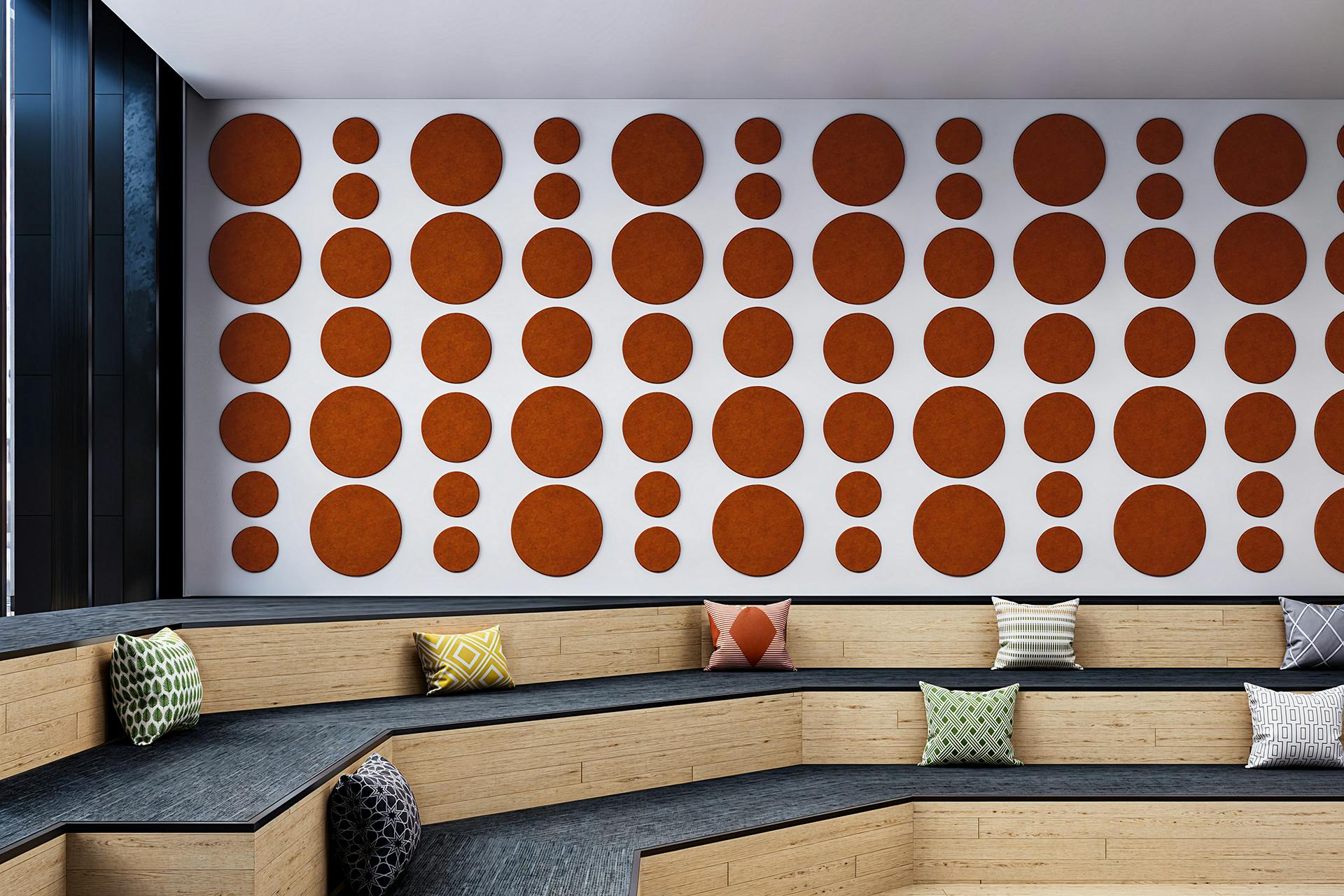 A modern interior space features a stepped wooden seating area with patterned cushions. The white wall in the background is decorated with numerous circular orange acoustic felt panels of various sizes, arranged in a symmetrical pattern. Black window frames are visible on the left.