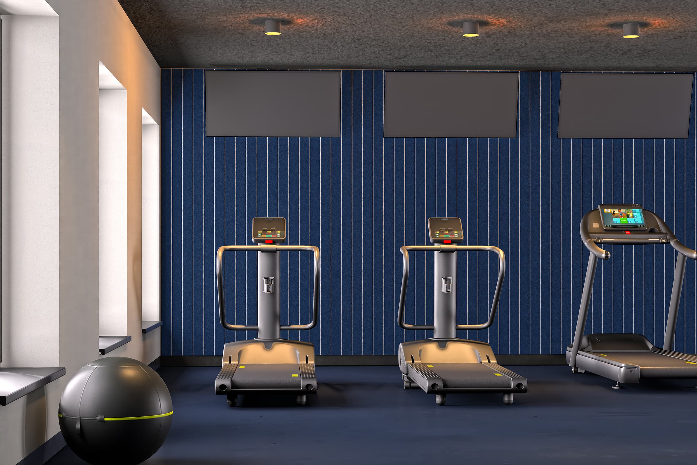 A modern gym room with three treadmills facing a blue striped acoustic felt wall, each with monitoring screens. There are three windows on the left, allowing natural light to enter. A large exercise ball is in the front left corner of the room. The floor is dark blue.