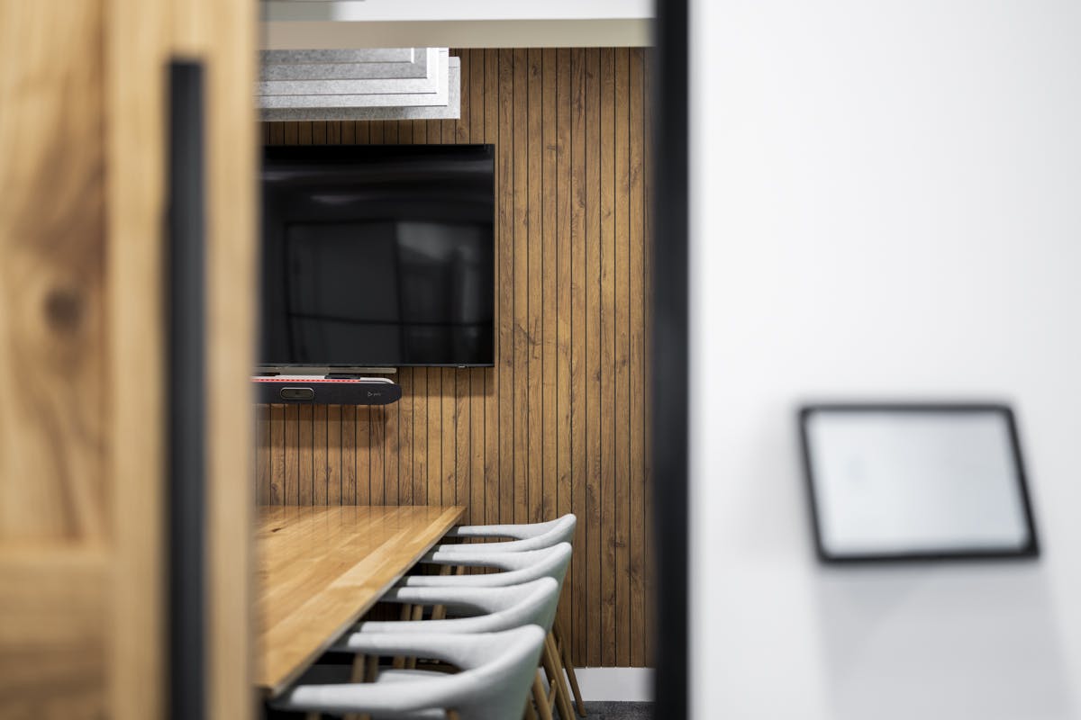 A modern meeting room in the GumGum Head Office with wood-printed acoustic felt walls, featuring a large flat-screen TV, a long wooden table, and several light gray chairs. A wall-mounted device is near the entrance. The room is partially visible through an open door.