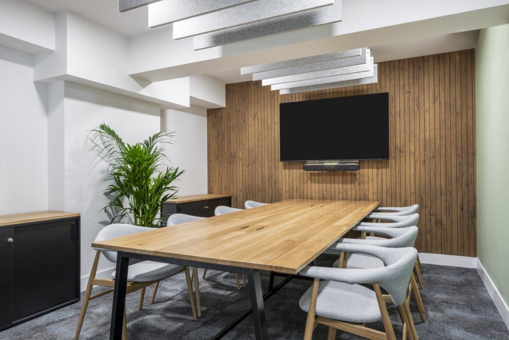 A modern conference room in the GumGum head office boasts a large wooden table surrounded by eight gray chairs. The room features a wood-paneled acoustic felt wall with a flat-screen TV mounted on it. There's a potted plant in the corner and acoustic ceiling panels hanging above the table.