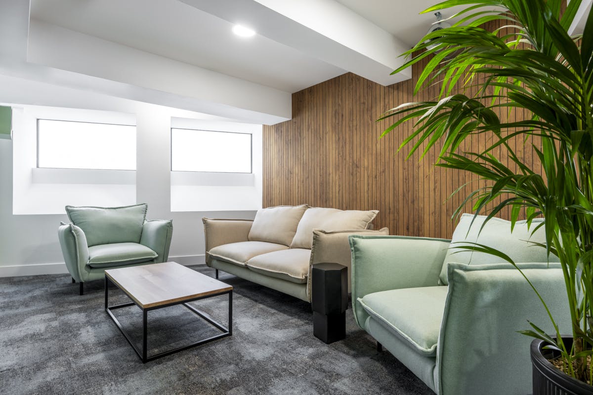 A modern lounge area in the GumGum head office features two light green armchairs, a beige sofa, a wooden coffee table, and a tall potted plant in the corner. The room boasts large windows and a paneled acoustic felt accent wall with gray carpeting on the floor.