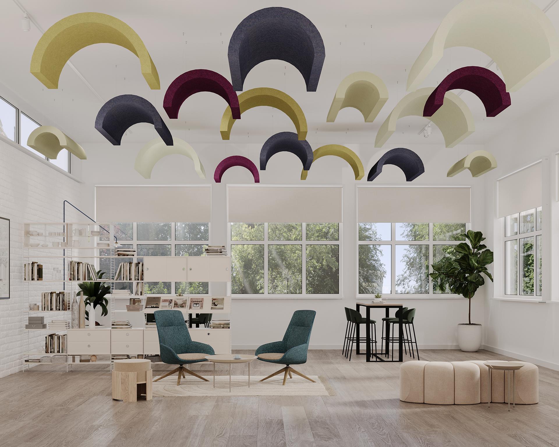 A bright, modern office space with large windows, light-colored floors, and white walls. Colorful curved acoustic felt clouds hang from the ceiling. The room features teal chairs, a wooden coffee table, bookshelves, an indoor plant, and a set of poufs and a table.