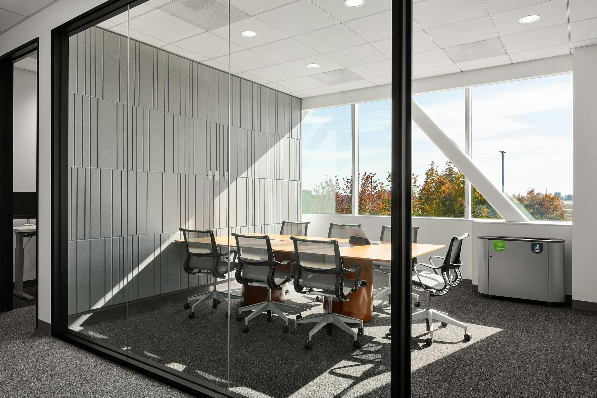 A modern conference room with a large wooden table surrounded by eight office chairs. The room features floor-to-ceiling windows allowing natural light to flood in, gray acoustic felt wall panels on one side, and a view of trees outside. A recycling bin stands near the windows.