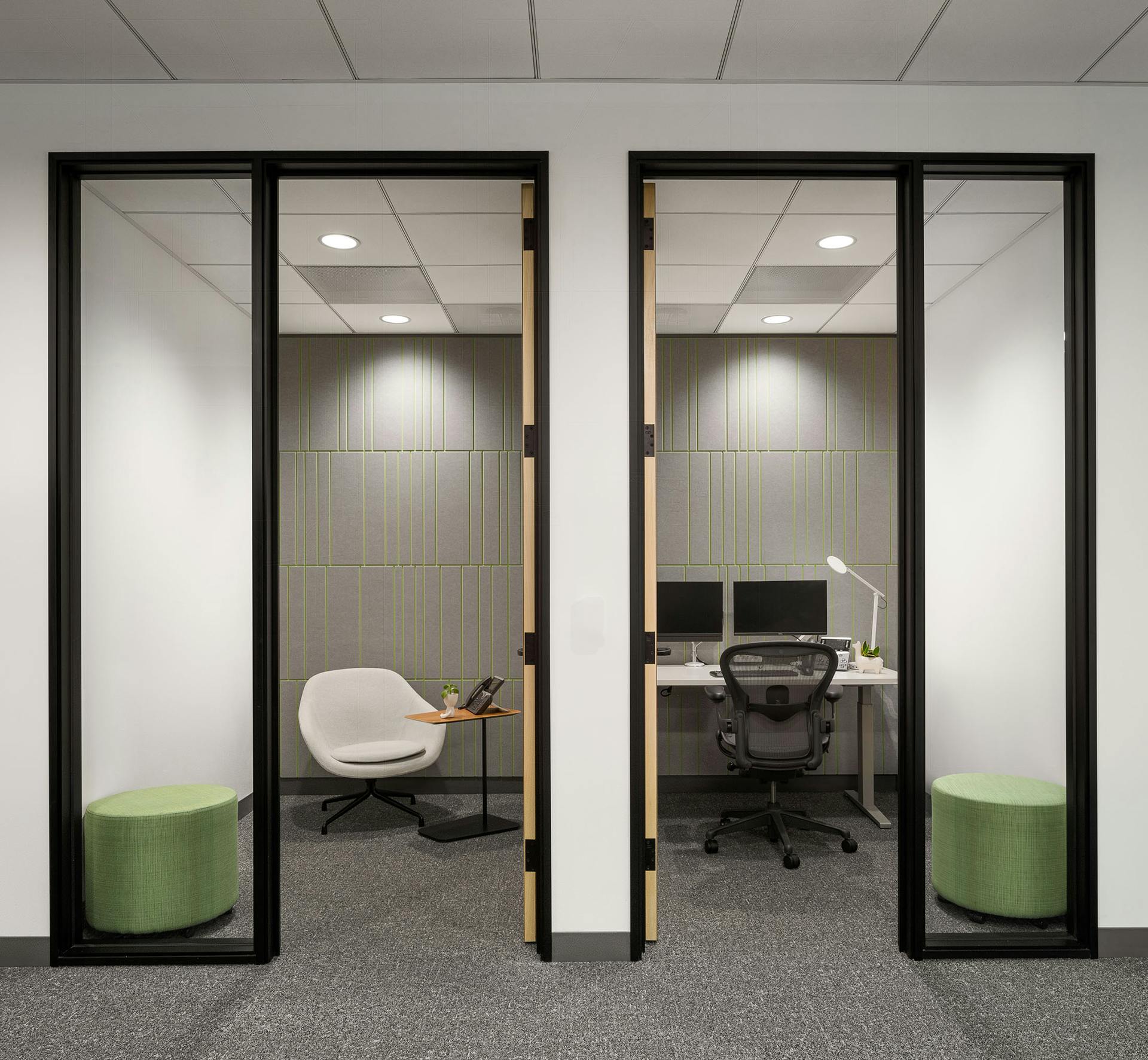 Two small, modern office spaces divided by a glass panel. Each office has a door with a tall window. The left office contains a white chair and green ottoman. The right office is equipped with a desk, task chair, dual monitors, keyboard, mouse, desk lamps. Both offices feature an acoustic felt wall panel for sound absorption.