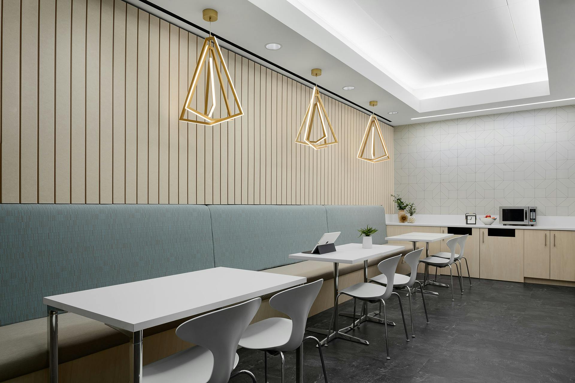 A modern break room with a minimalist design features white tables and gray chairs. Golden geometric pendant lights hang above a light blue cushioned bench. The floor is dark, and the walls boast an acoustic felt wall panel and a light patterned section. Kitchen amenities are visible in the background.