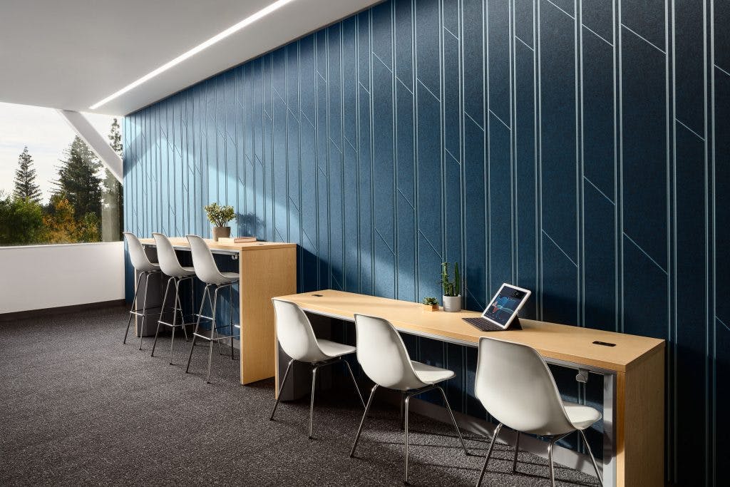 A modern interior space features a long wooden counter with white chairs lined up against a blue wall adorned with geometric patterns in acoustic felt wall panels. On the counter, there are plants and a tablet. Bar stools are placed at another counter by a window with a view of the outdoors.