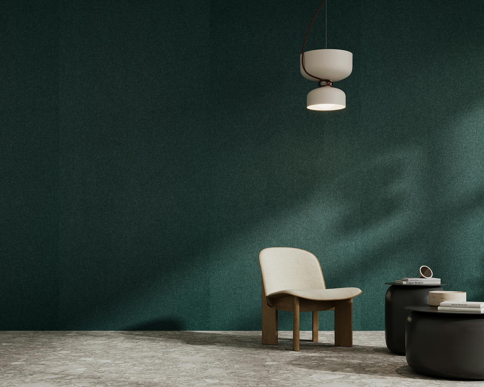 A minimalist room features a single light wooden chair with a white cushion, positioned next to two black round side tables. A modern white pendant light hangs from the ceiling, casting a soft glow. The walls are adorned with dark green acoustic felt panels and the floor is stone-textured.