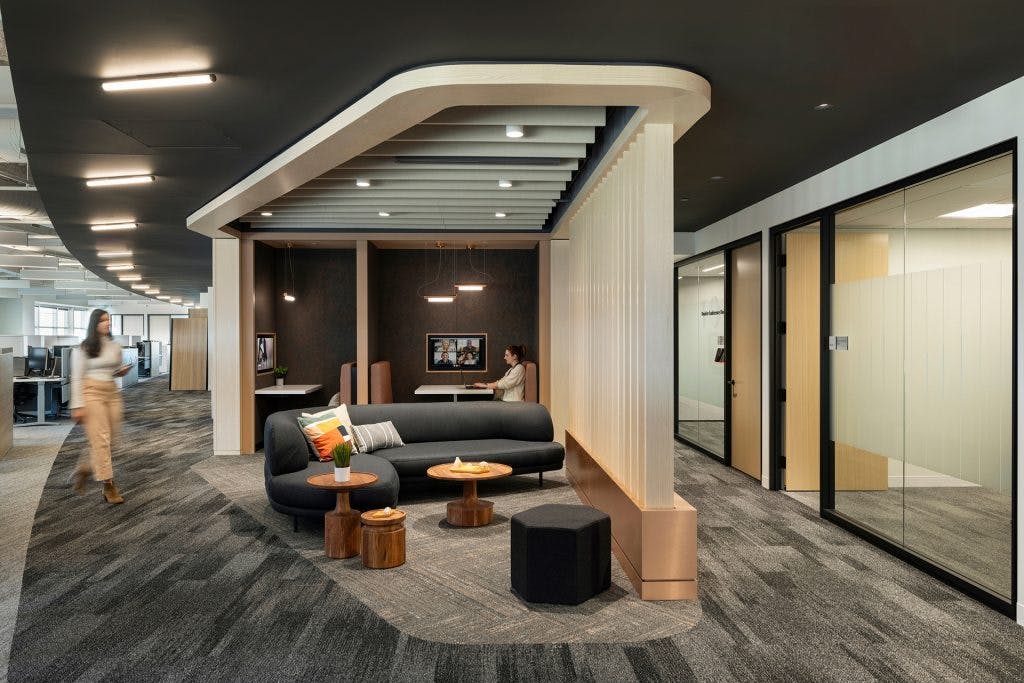 Modern office with sleek design features a small seating area with a black sofa, wooden tables, and hexagonal ottomans. Acoustic ceiling baffles enhance the space's acoustics while providing a stylish touch. A person works on a laptop at a booth in the background, with glass-walled conference rooms and workstations visible throughout the open space.