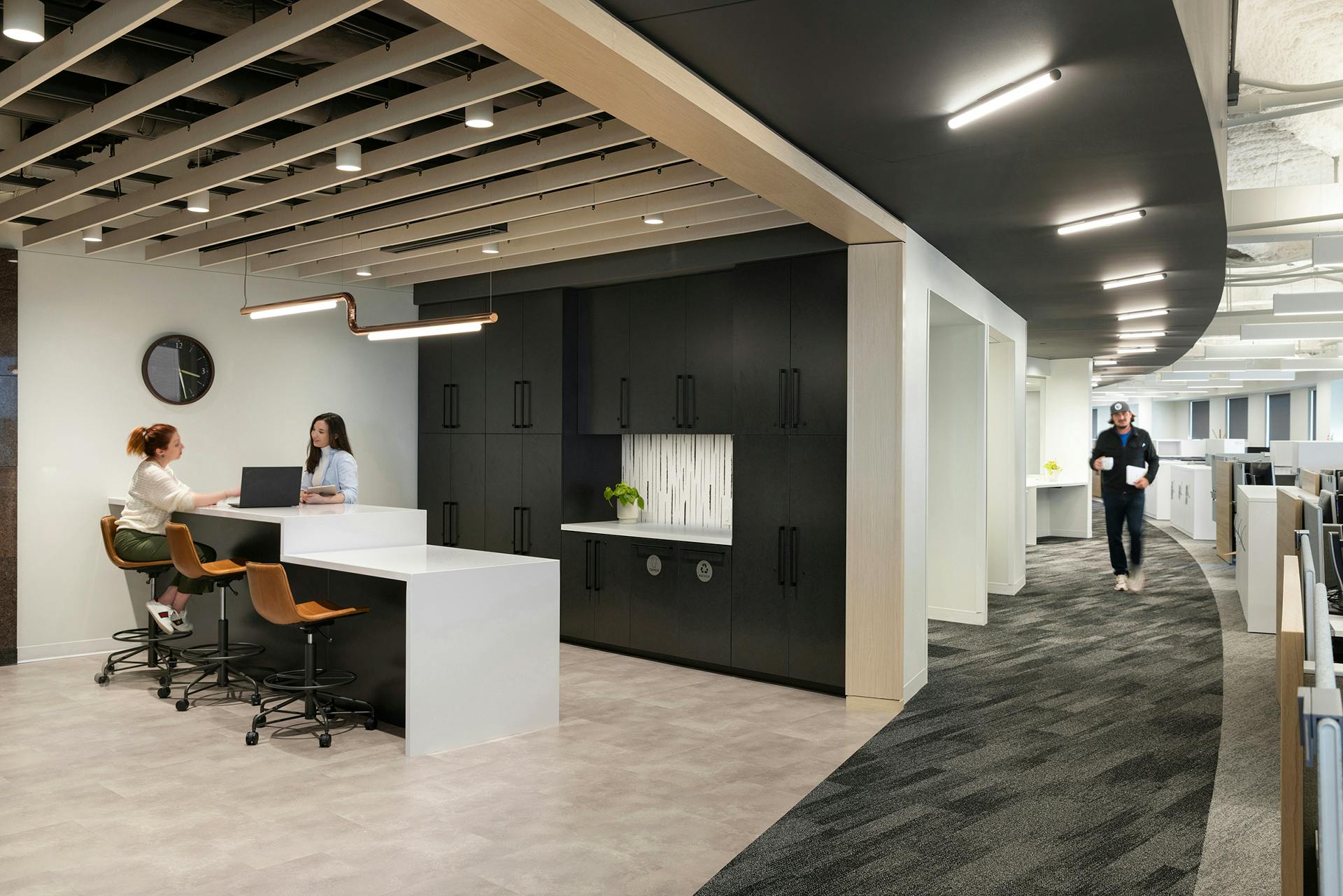 A modern office kitchen area with light wooden floors and white countertops. Two women are sitting on stools at a counter, engaged in conversation under an acoustic ceiling baffle. A man is walking through a corridor with cubicles to the right. Dark cabinets and a clock are on the wall in the background.