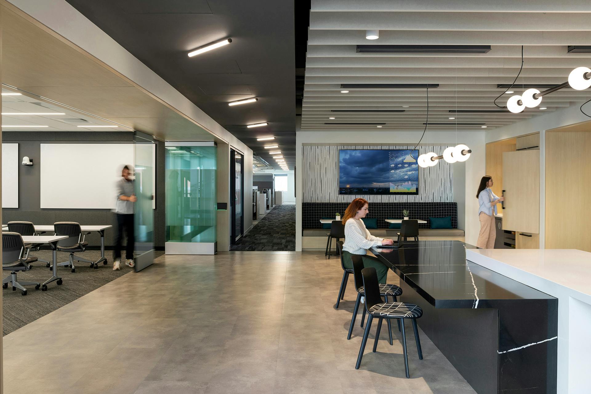 Modern office space with various working areas. A woman types on a laptop at a high table in the foreground, while another person stands near a TV screen in a conference room. An individual walks down a hallway, beneath stylish acoustic ceiling baffles. Overhead lighting and sleek furnishings are visible.