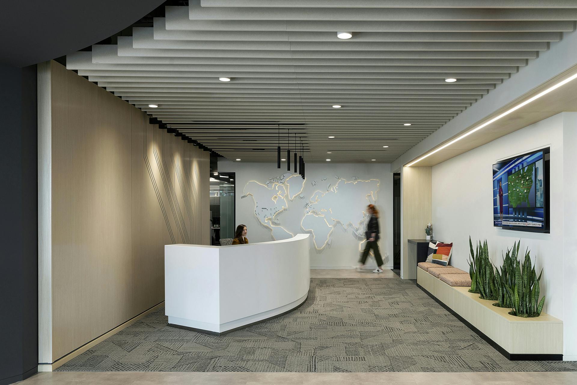 A modern office reception area with a curved white desk staffed by a seated person. A map of the world is mounted on the wall behind. A person walks by. Seating with cushions and plants is on one side, and a flat-screen TV is mounted on the wall. The ceiling features acoustic ceiling baffles for sound quality.