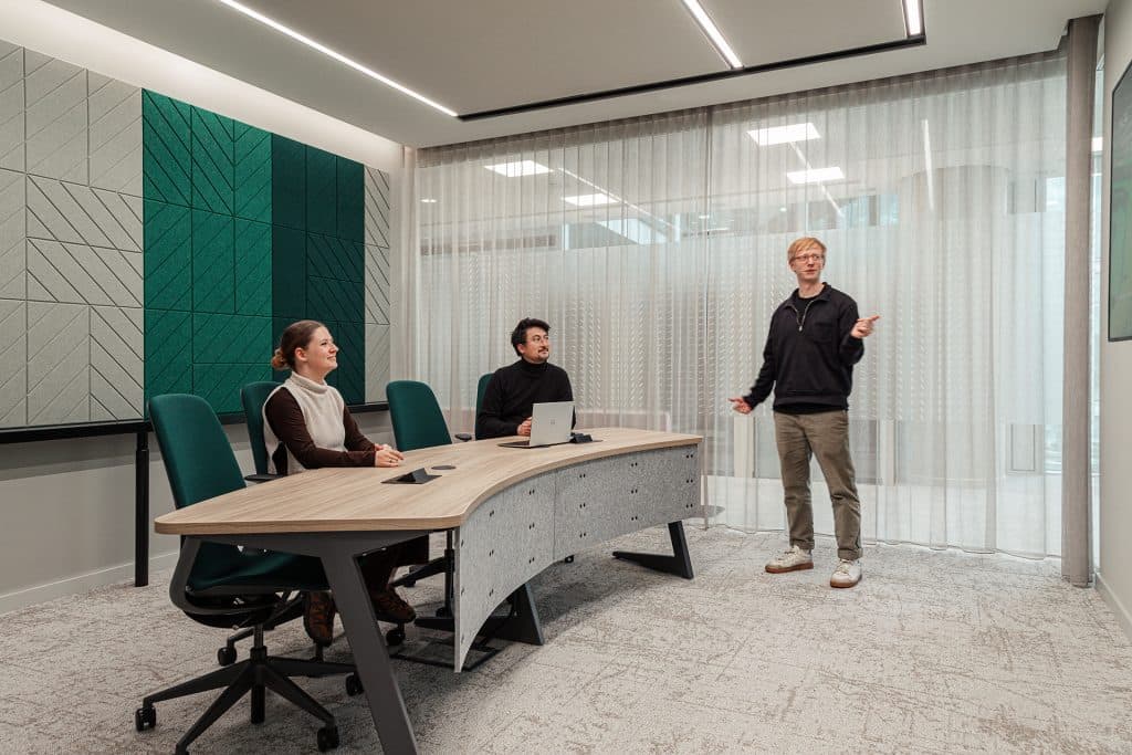 A man stands and gestures towards a screen in a modern conference room, while two seated colleagues, a woman and a man, attentively listen. The room features contemporary decor with geometric acoustic felt wall panels, a large table, and ample natural light filtering through sheer curtains.