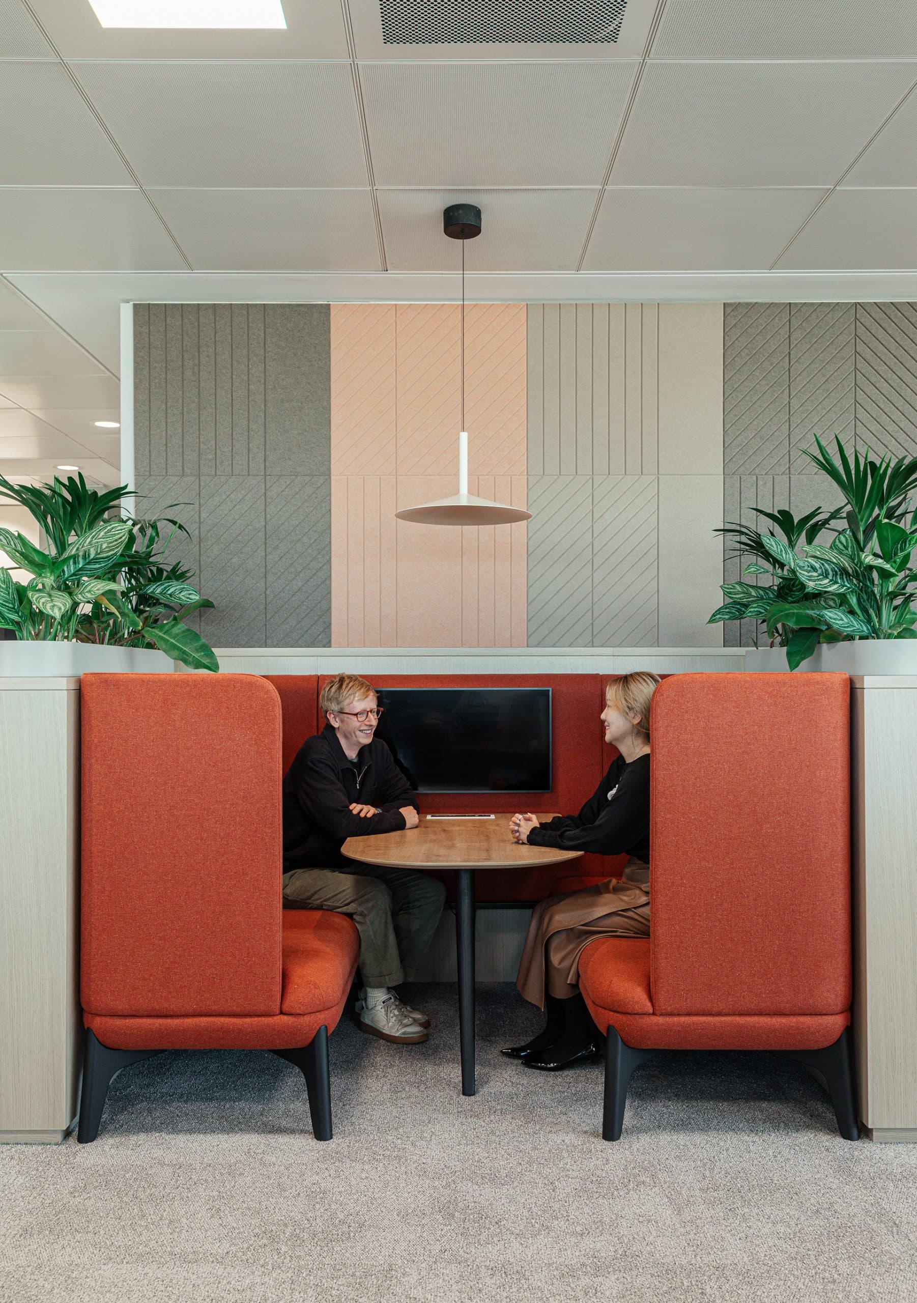 Two people seated across from each other in a semi-enclosed booth with high orange seats in a modern office setting. The booth has a small wooden table between them and a screen on the wall. Green plants flank both sides of the booth. Geometric acoustic wall panels are visible.