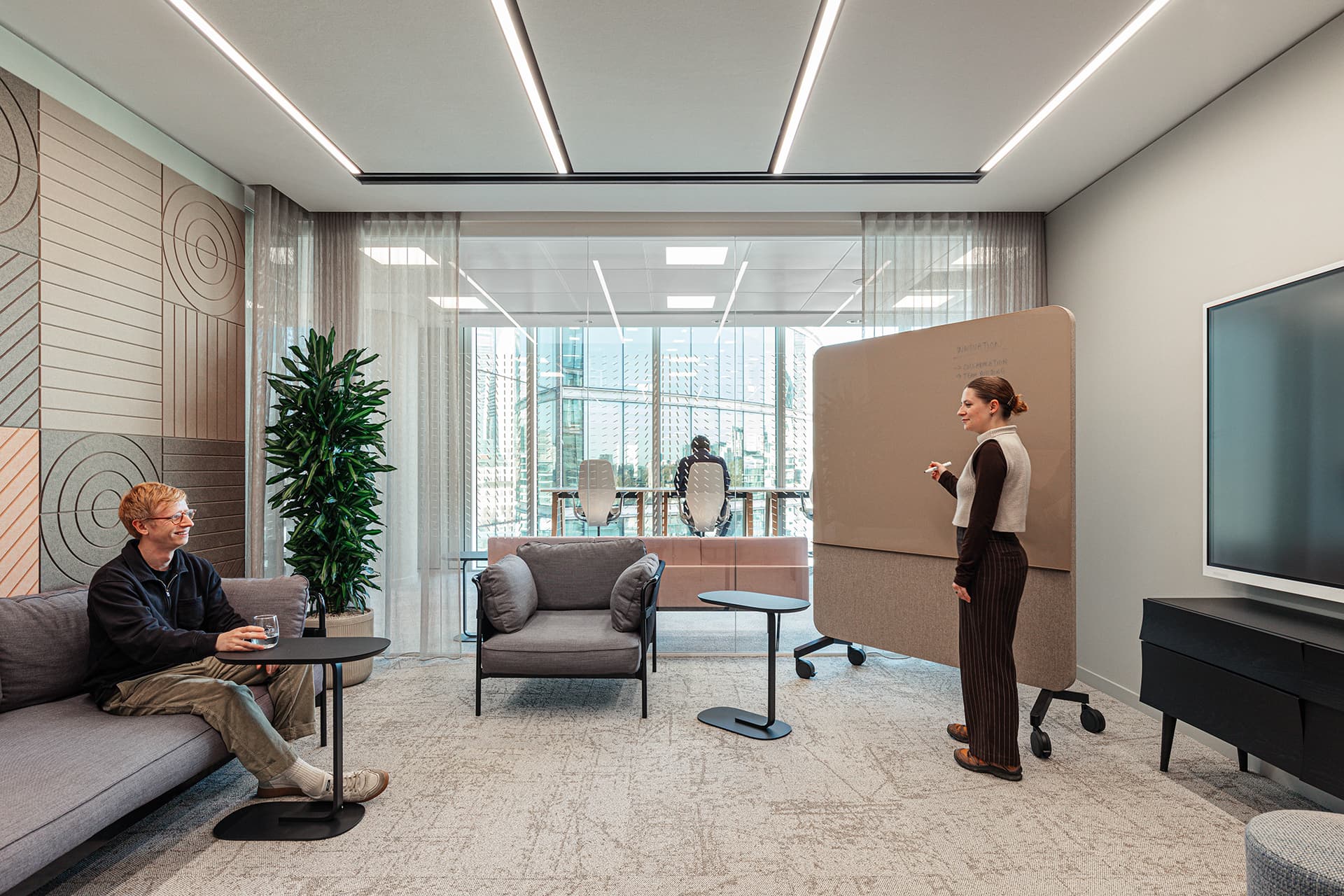 A modern office space with two people. One person is seated on a couch with a drink, while the other stands writing on a mobile whiteboard. The room features stylish decor, an acoustic felt wall with geometric patterns, a large window with a city view, a plant, and contemporary furniture, including a flat-screen TV.