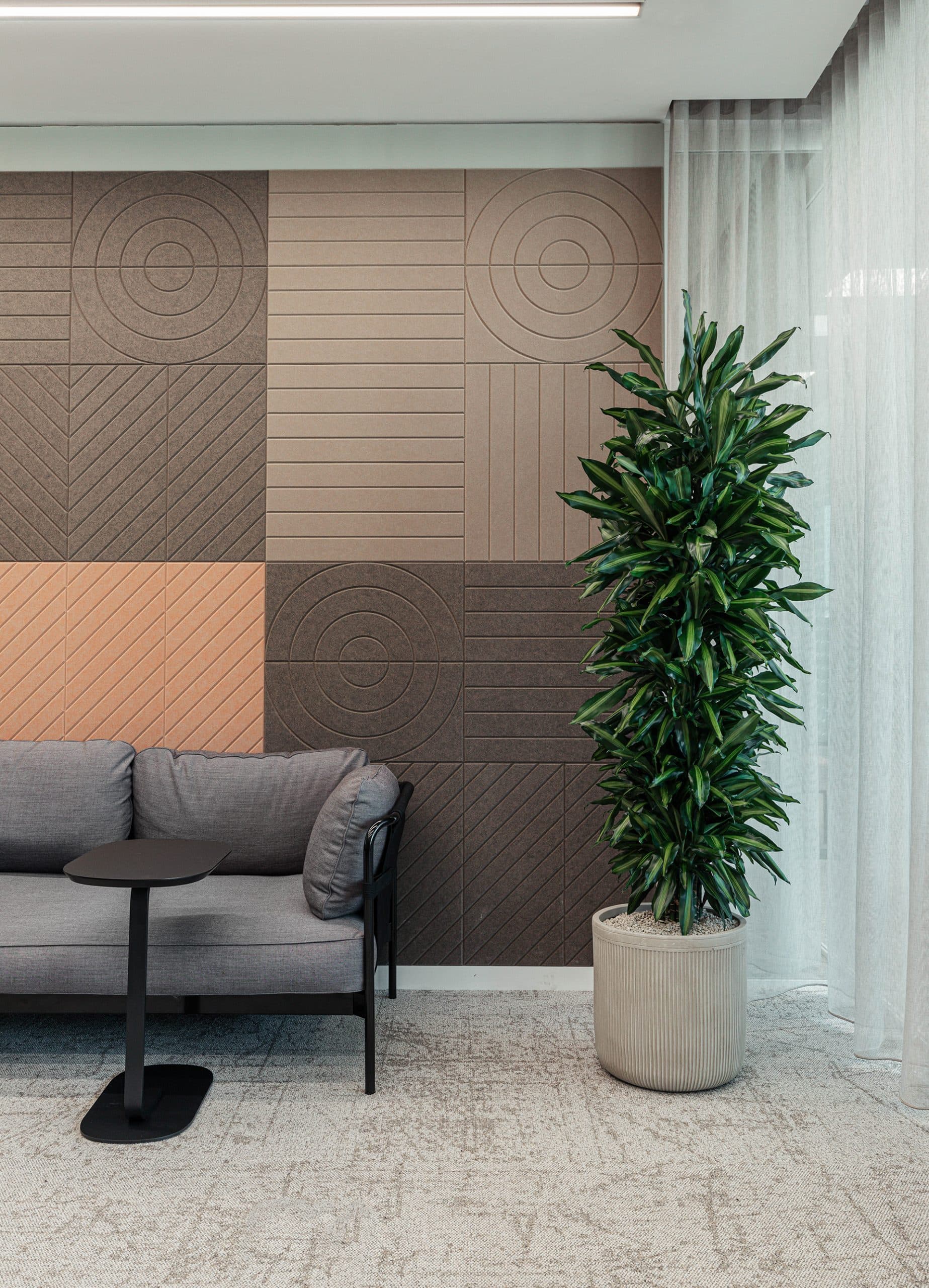 A modern office space with a grey cushioned sofa on the left, a small black table beside it, and a tall potted plant with green leaves on the right. The background features an acoustic felt wall with geometric patterns and a floor covered with light-colored carpet.