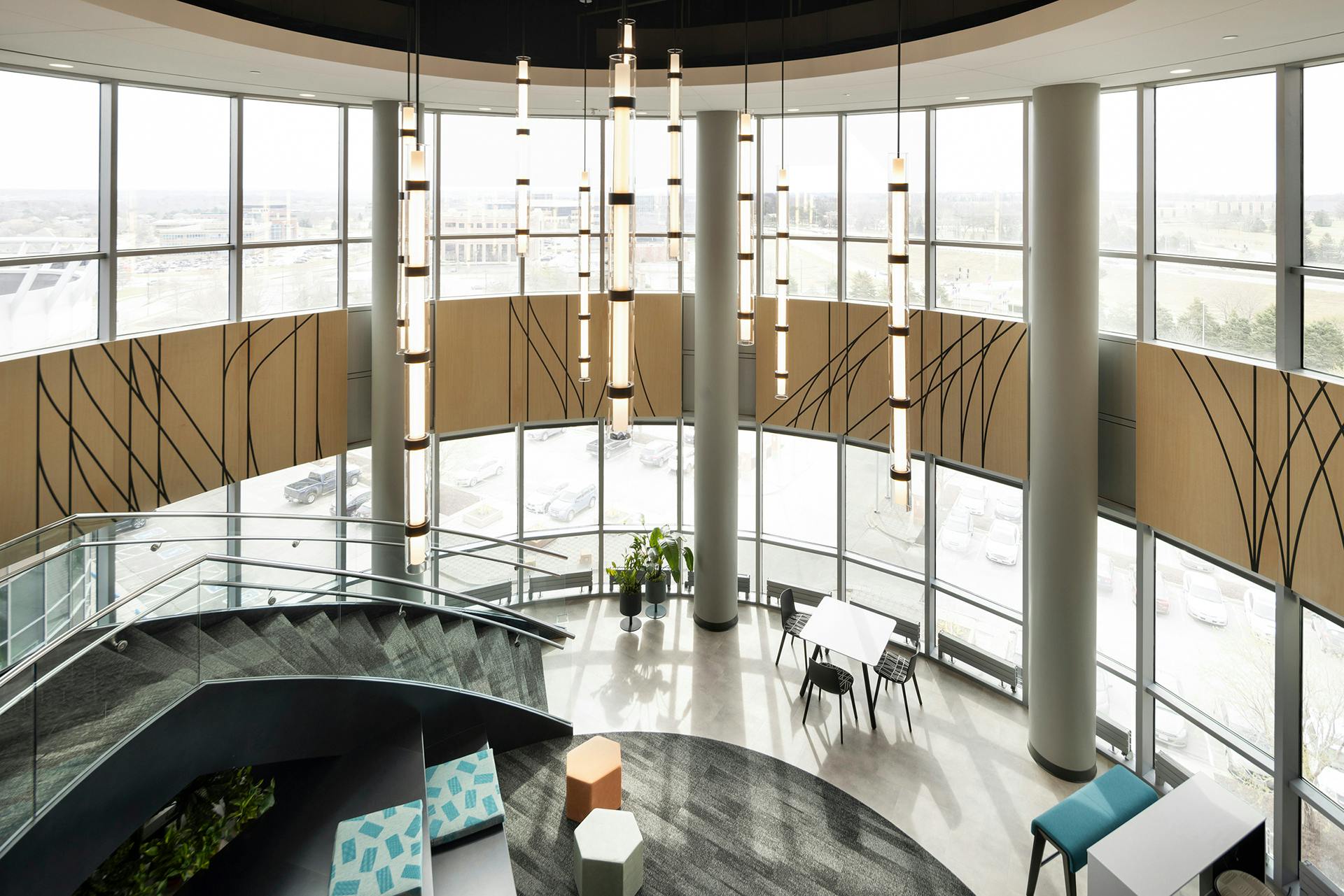 A modern, open-concept office space with floor-to-ceiling windows, curved staircases, and pendant lights. The design includes geometric acoustic felt wall patterns, diverse seating options, and a bright, airy atmosphere with natural light flooding in.