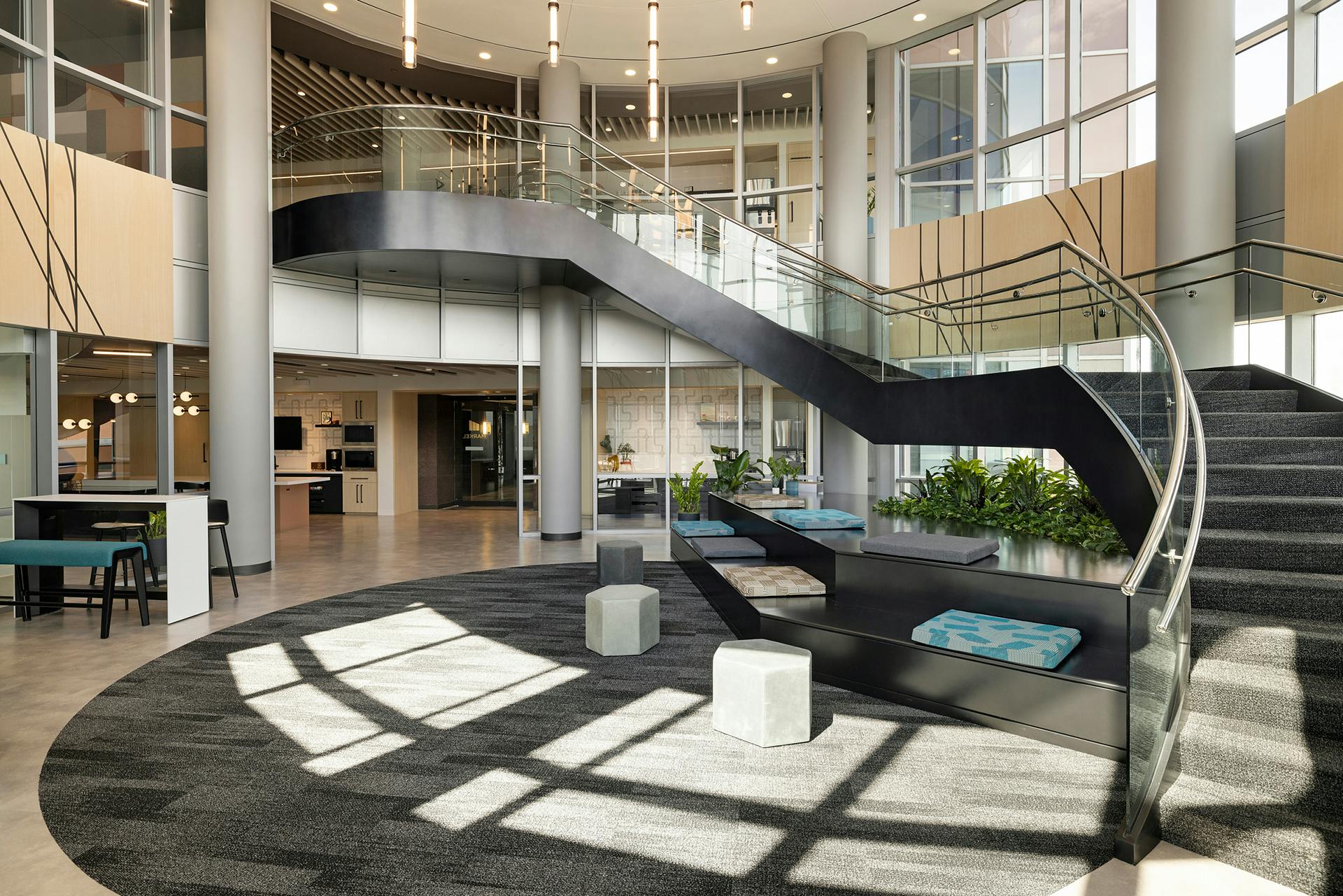 A modern lobby with a curved staircase, large windows, and plenty of natural light. There are seating areas with green cushions around the staircase, a few square stools, and indoor plants. The space features acoustic felt wall panels alongside a mix of carpet and tile flooring for a light and open design.