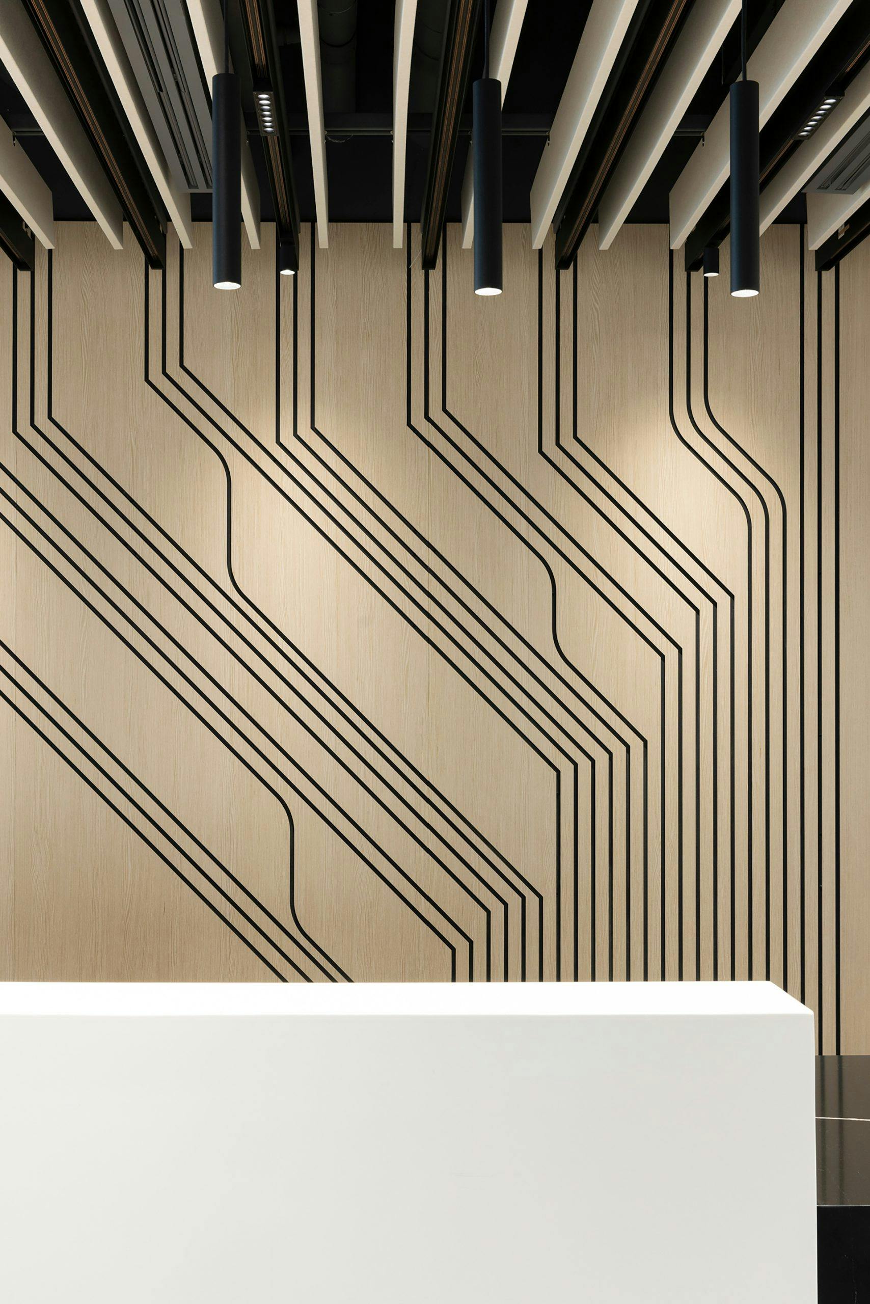 A modern, minimalist interior featuring an acoustic felt wall panel with black geometric lines forming an abstract pattern. The ceiling has black-and-white beams with cylindrical pendant lights. A white reception desk sits in the foreground.