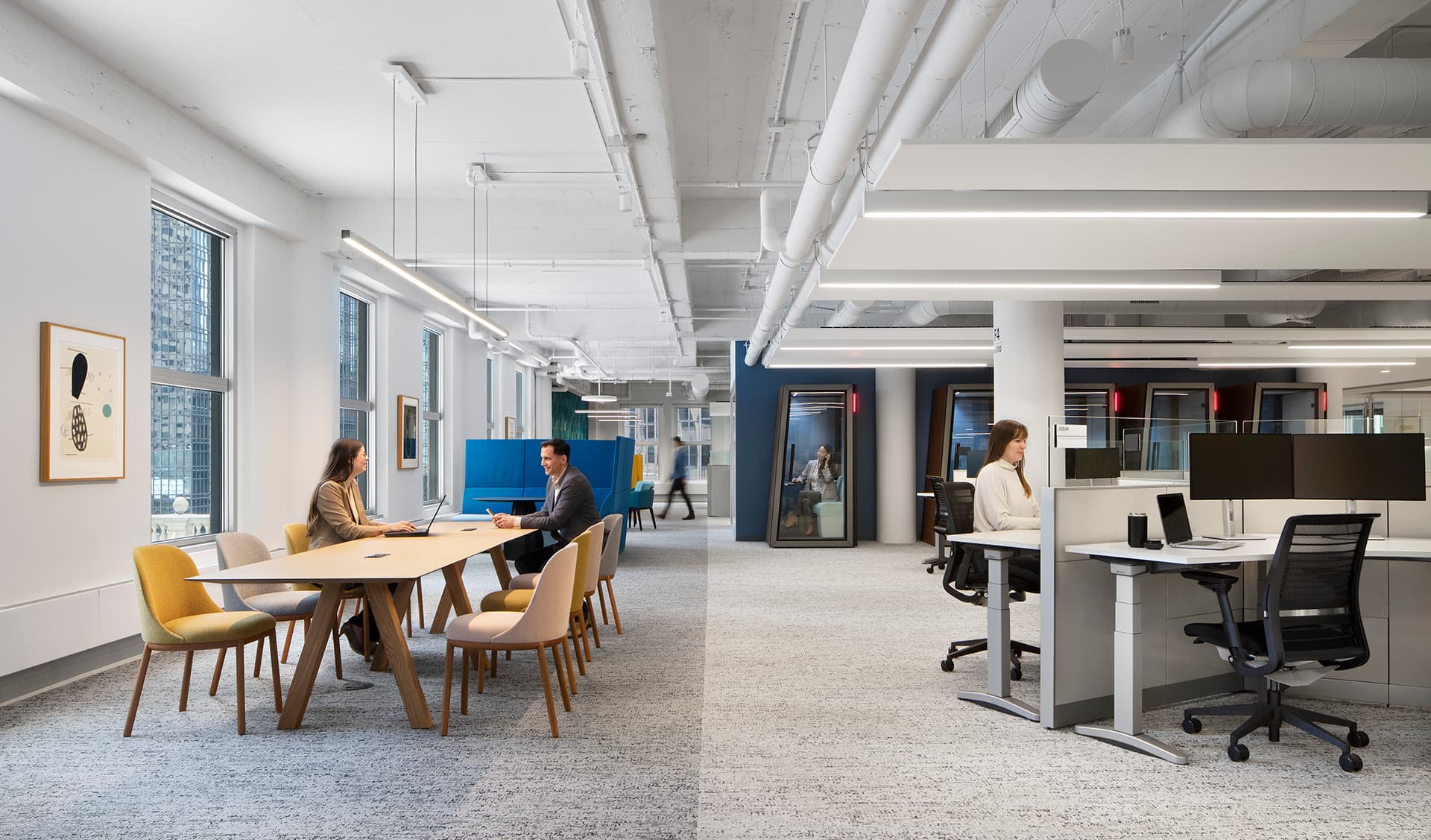 A modern office in Minnesota with white ceilings and walls. Two people discuss finance at a round table while another works at a desk on a computer. Natural light streams through large windows, and contemporary artwork decorates the walls, creating an inspiring workspace.