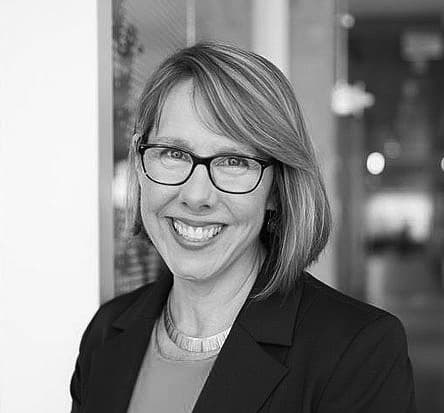 A person with glasses and shoulder-length hair smiling while wearing a suit jacket and a necklace. They stand indoors with a blurred background, conveying a professional setting. The image is in black and white.